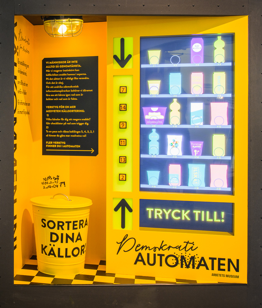 En del av utställningen Välkommen in! – Demokrati för vuxna, byggd som en försäljningsautomat i gult och svart. Intill automaten står en soptunna med texten "Sortera dina källor".