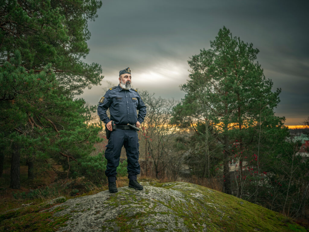 En polisman i uniform står på en bergknalle med händerna i sidorna. Han är omgärdad av tallar, bebyggelse anas i bakgrunden.