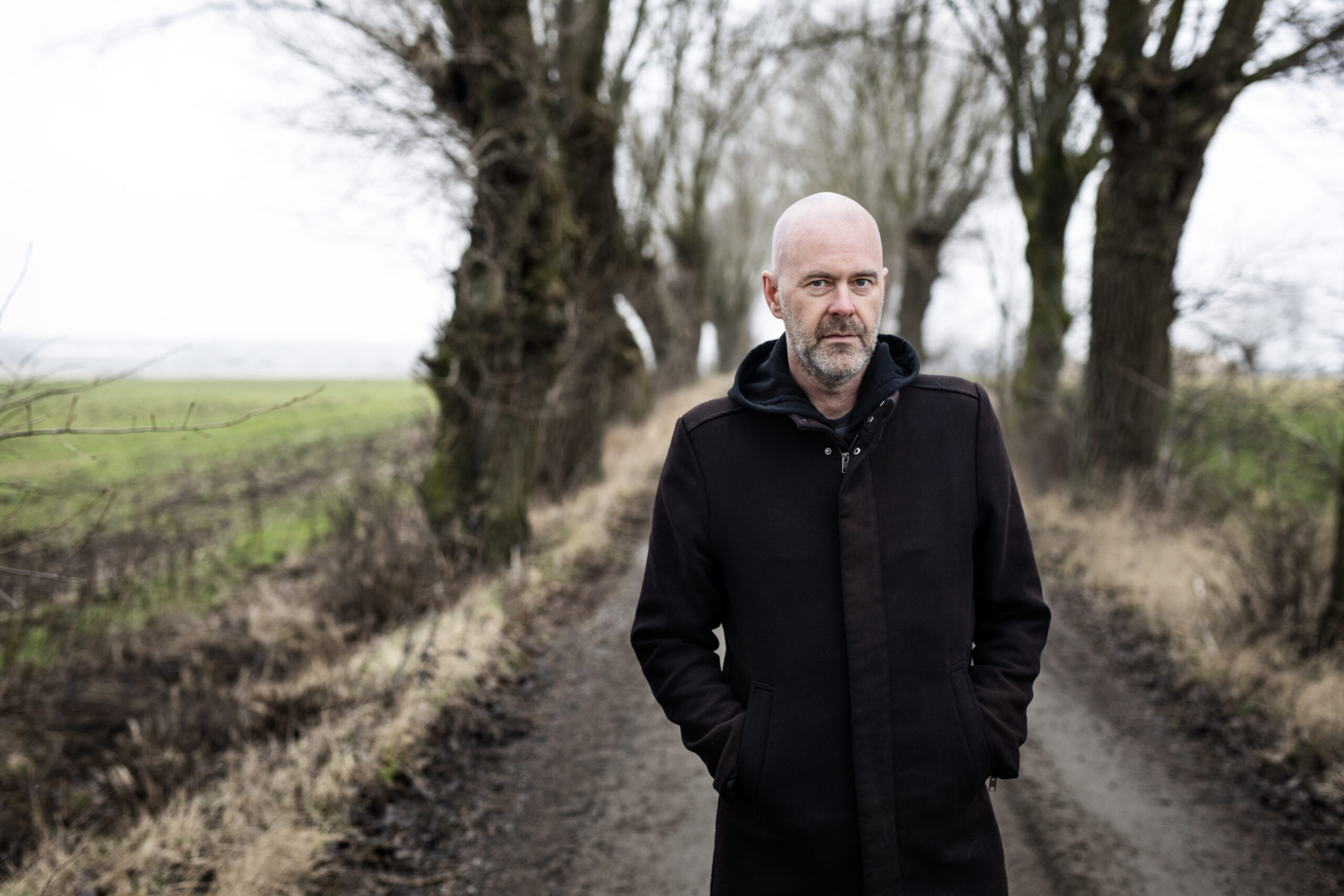Författaren Patrik Svensson i en svart rock. Han står på en grusväg i en allé och vädret är grått och dimmigt.