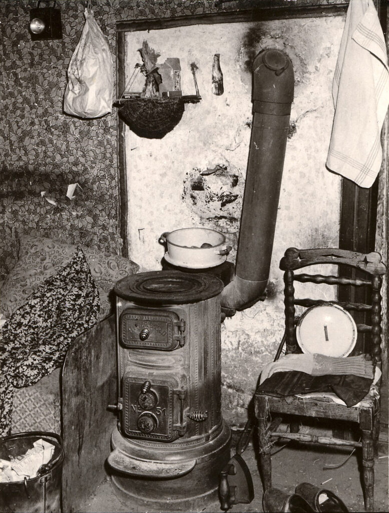 Svartvitt foto. Bild inifrån ett statarhem. I mitten syns en kamin i gjutjärn, med en spisplatta. Runtom syns kanten av en säng, en hink, en kastrull, en trästol med ett kastrullock och ett par sockar på, ett strykjärn och ett par träskor. På väggen hänger en påse, en liten hylla och en kökshandduk. Väggen består av en vit spismur och småblommig tapet, med sotfläckar. 