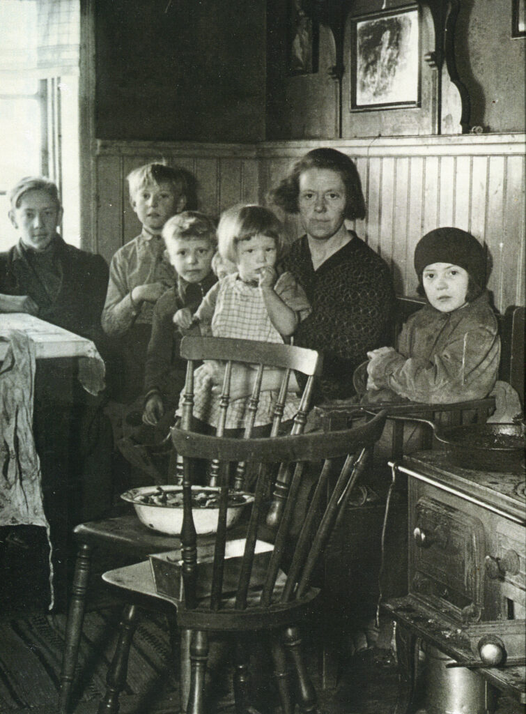 Svartvitt foto. En kvinna med fem barn sitter i en kökssoffa, inne i en statarbostad. I bilden syns köksbordet, en järnspis och två stolar. På stolarna står en skål och en plåtlåda. På spisen står en stekpanna. Väggen är delvis klädd i panel och en tavla. På golvet ligger en trasmatta.