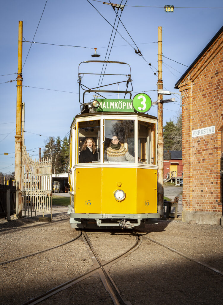 En gul spårvagn med två kvinnor i förarhytten rullar fram i industrimiljö mot en klarblå himmel.